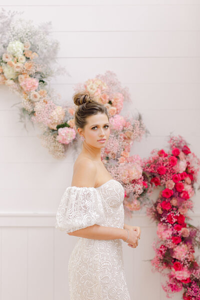 Woman in white lace off the shoulder dress in front of a white wall with a curving floral arrangement of gradient pink colors