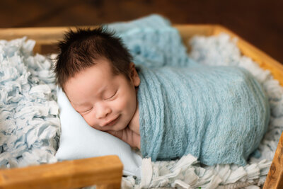 Newborn photographer, a newborn baby in a blue swaddles smiles while lying on a bed.