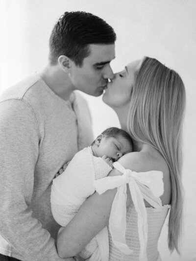 mom and dad kissing with newborn baby asleep on shoulder in black and white photo by seattle family photographer jacqueline benet