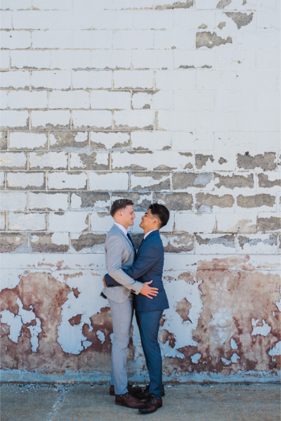 Two Grooms in black and gray suits holding each other staring into each other’s eyes in front of a brick wall.