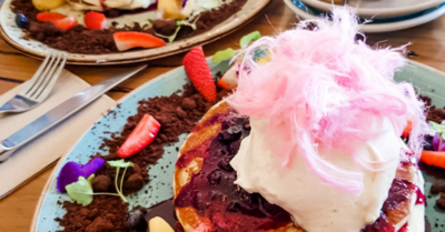 An image of blueberry pancakes, cream, flowers, chocolate, strawberries and fairy floss. There are two servings of pancakes in the image; one is on an aqua plate and the other plate is white.