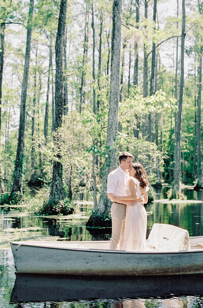 Engagement session on a sailboat. She is wearing a white halter top swimming suit sitting on the side of the boat with one leg down towards the water and the other one lifted straight out as she is kicking her legs and looking up at him. He is wearing white shorts with a white short sleeved dress shirt, standing behind her looking down at her while holding on the the rope of the sail. You can see water and a bridge behind them. Photographed by wedding photographers in Charleston  Amy Mulder Photography