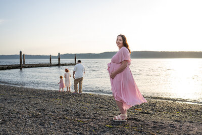 a pregnant mother is standing at the beach with her husband and kids playing in the water behind her