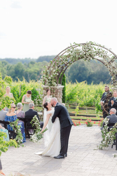 Bride and groom share a romantic kiss under a floral arch at their outdoor vineyard wedding ceremony, captured by Destination Wedding Photographer.