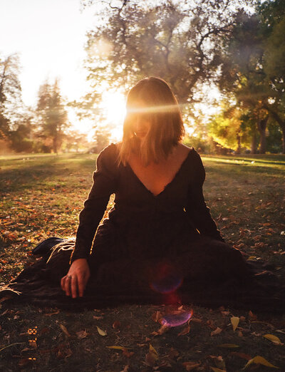 A serene outdoor image of a woman seated on the ground in a park during golden hour. The sunlight streams through the trees, creating a warm glow and lens flares that partially obscure her face. She wears a dark dress and appears peaceful, surrounded by autumn leaves scattered on the grass. The background is filled with soft-focus trees and warm light, enhancing the tranquil atmosphere.