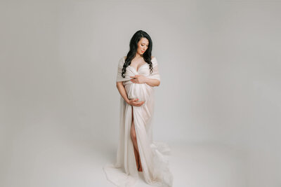 pregnant mom with black hair wrapped in white silk on white background holding pregnant belly