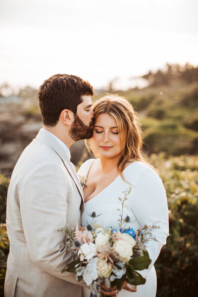 Groom kissing bride’s forehead as she holds a bouquet on the rocky coast during their Maine wedding.
