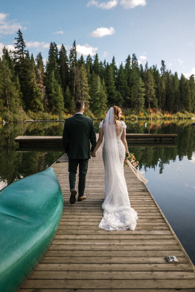 A bride and groom hold hands facing away from the camera. They are standing on a dock with a canoe in the foreground and a lake in the background.