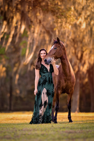Bay horse nuzzles equestrian wearing a long green dress under a sunlit tree.