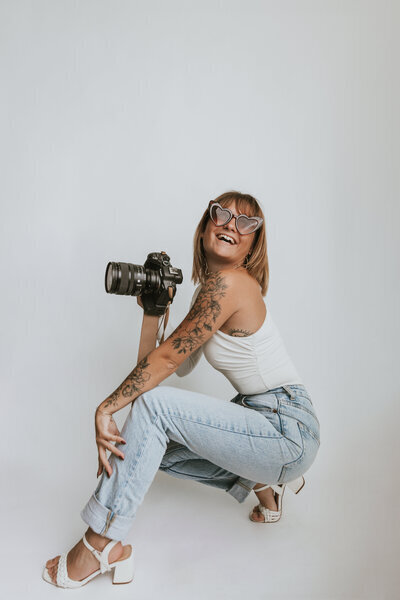 Laura, photographe mariage, de profil, accroupie avec son appareil photo, lunettes en forme de cœur, tatouages apparents, jean et haut blanc.