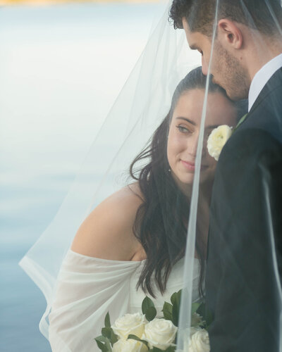 A bride and groom hug under a veil, the bride looks off camera and the groom kisses her on the forehead during portraits after their catholic wedding mass at St. margaret Mary's mission church in Decherd, Tennessee