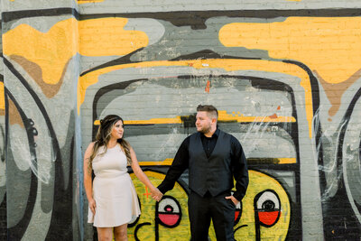 Engaged couple hold hands and look at one another in front a mural painted wall
