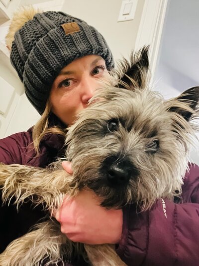 blonde woman in beanie kissing a grey cairn terrier dog in her arms