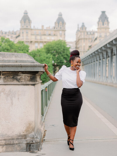 a black woman in a white top and black skirt smiling and looking off into the direction of the bir hakeim bridge in paris
