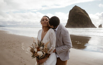 Bride holding a dried floral bouquet as the groom kisses on the Oregon coast.