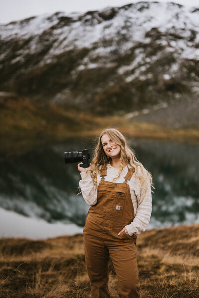 a photographer smiling at the camera wearing carhartt overalls in hatcher pass with a lake and mountains behind her