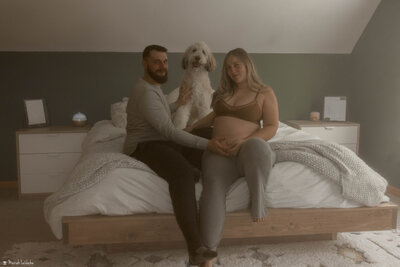 couples sitting on bed with dog holding pregnant belly