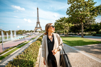 A woman wearing sunglasses and a light jacket stands smiling in a park with the Eiffel Tower in the background. The scene is sunny, with fountains and trees lining the path.