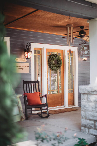 Welcoming entrance to 'The Porch' counseling center featuring a wooden door with glass panels flanked by sidelights, adorned with a fresh green wreath. A black rocking chair with a bright orange cushion sits on the covered porch. Gray horizontal siding, wall-mounted lantern sconces, and a wooden beam ceiling create a warm, inviting atmosphere. A 'Welcome' sign and wind chimes complete the cozy, peaceful setting.