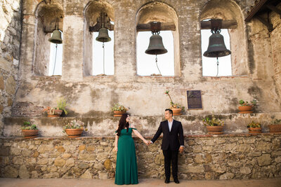 Engaged couple gaze at each other while holding hands under the bells in the bell courtyard area at the Mission San Juan Capistrano