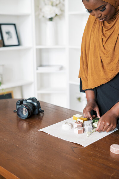 Woman wearing burnt yellow hijab adjusting ring boxes on fabric with Canon camera sitting to the left
