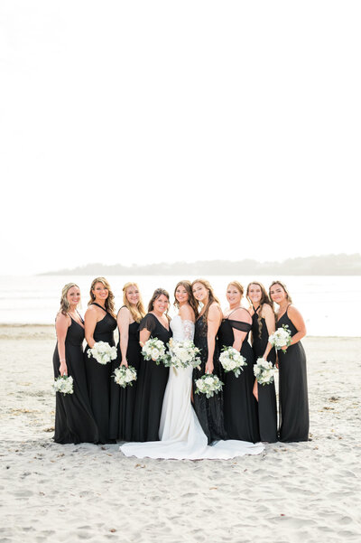A bridal party stands on the sandy shore at Newport Beach House, with the bride in a fitted lace gown and bridesmaids in elegant black dresses holding white rose bouquets.