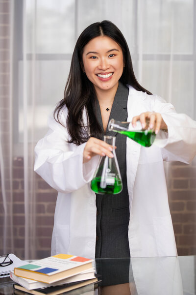 Stephy Chen smiling in a white lab coat while holding a beaker during a creative personal brand photoshoot in a modern office.