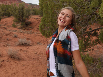 Blonde haired woman smiling in front of a tree, wearing a tribal scarf and a white shirt.