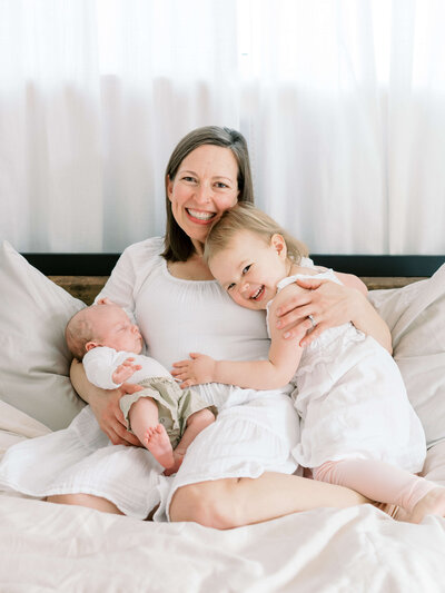 New mom sits on the bed and hugs her toddler daughter and her baby son during a family photography session