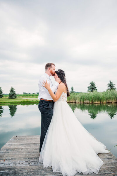 A groom kisses the bride's forehead as she has her hands around him. they are standing in front of a lake with evergreen trees in the background.