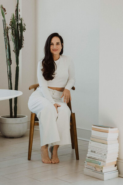 Ayurvedic Skin Specialist, Jen, wearing a white top and pants sitting on a jute rug leaning against a neutral couch