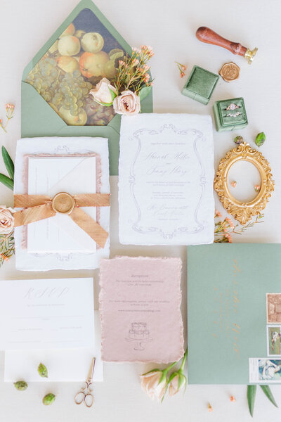 A stunning flat lay including white, dusty rose, and sage green stationary  with gold wedding rings, pink and green florals, and a velvet green ring box