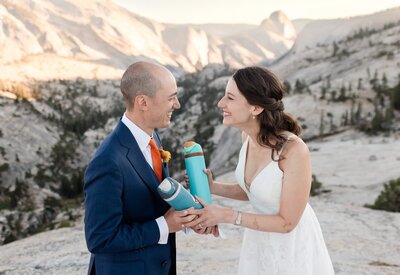 A bride and groom laugh while placing their custom wedding date stickers on their water bottles, while standing in view of Half Dome in Yosemite National Park. 