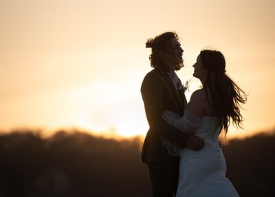 A bride and groom embrace after private vows during their luxury adventure elopement on a hidden beach in Mexico.  They are silhouetted by the glowing orange and yellow sunset that fills the sky.  They both have long hair, and it is blowing in the wind.  