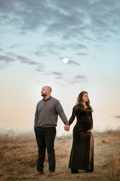 Couple stand in a field at twilight while the mother holds her pregnant belly. 