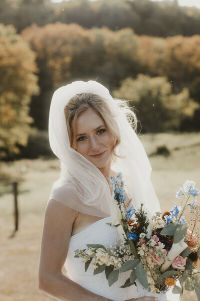 A smiling bride wearing a white veil and strapless gown holds a colorful bouquet of wildflowers, standing outdoors with trees and sunlight in the background.