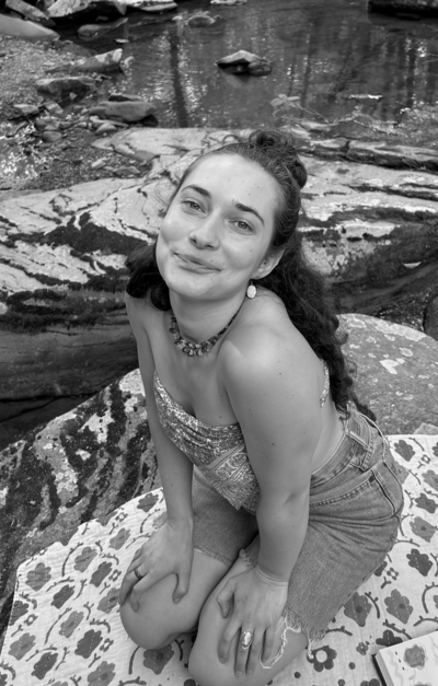 Black and white photo of woman with curly brown hair sitting on her knees by the river and softly smiling to the camera.
