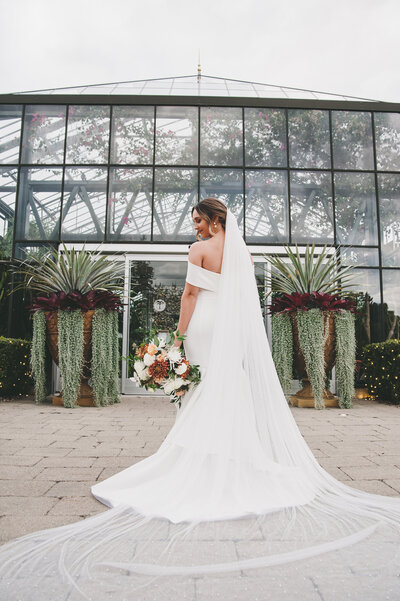 Alyssa Amez a custom wedding stationery and signage designer and business woman from Alyssa Amez Design as a bride on her wedding day at Planterra Conservatory in West Bloomfield, Michigan posing in her wedding dress and a flower bouquet in front of the wedding venue