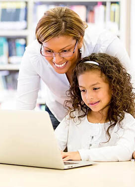 Female teacher assisting a young girl using a laptop inside a library.