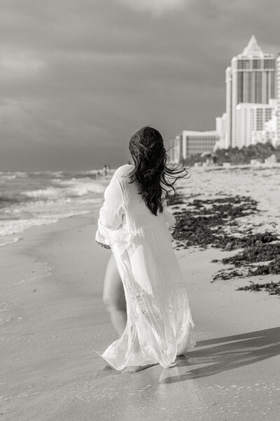 Pregnant woman in lace robe walking barefoot on Miami beach at sunrise, black and white photo.