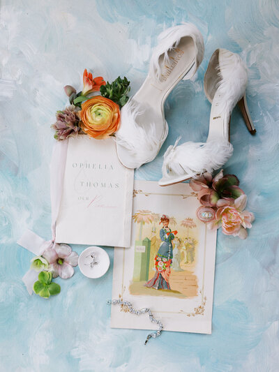bridal detail flat lay with invitations, feathered bridal heels, and florals on a blue hand-painted backdrop