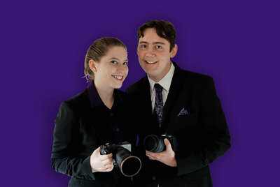 Zack and Shavonne, wedding photographers, posing with cameras in formal attire against a purple background.