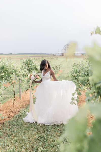 Bride holds bouquet and swings her wedding dress skirt amidst vines of a vineyard on her wedding day