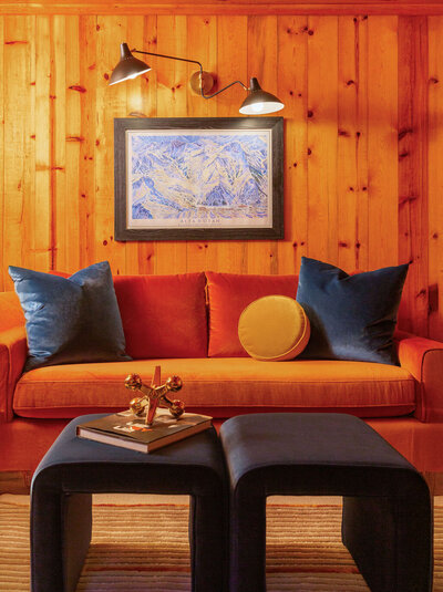 A warmly lit seating area in a wood cabin with orange-toned wood-paneled walls. An orange velvet sofa with rust-colored cushions is adorned with dark blue velvet and a round mustard yellow pillow. Two dark blue velvet ottomans sit in front, one holding a book and a gold geometric sculpture. Framed artwork hangs on the wall above the sofa, illuminated by two adjustable black wall sconces.