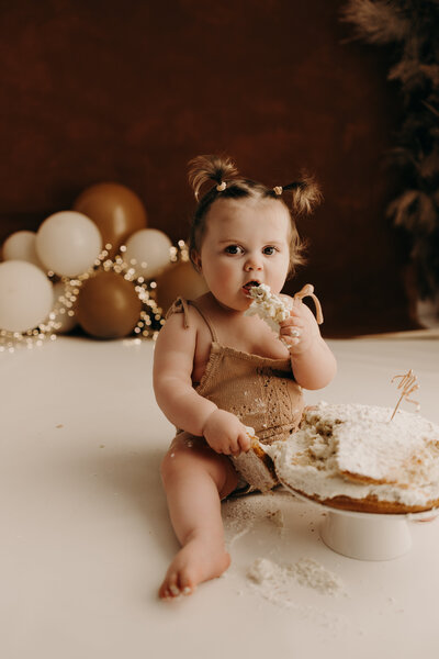 Baby girl celebrating first birthday with a cake and balloons, arms raised in excitement and surprise.
