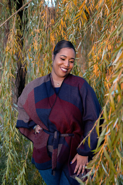 African American woman in red and blue sweater jacket, blue jeans in front of a weeping willow tree