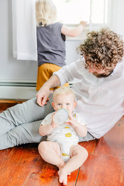 Dad looks down at his happy baby, who is holding a bottle, while another boy looks out the window in the background