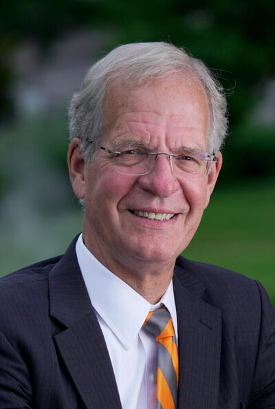 Smiling senior man wearing a dark pinstripe suit, white dress shirt, and orange-gray striped tie, posing outdoors with greenery in the background.