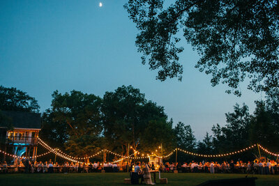 a bride and groom sit under a moonlit sky in front of all their wedding guests at long tables