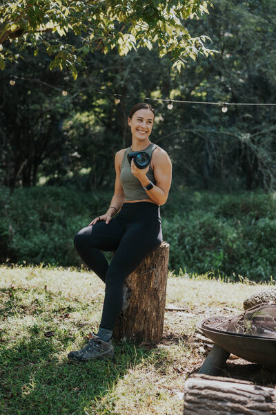 A photographer sits on a log while holding a camera and smiling off in the distance. She is surrounded by lush green grass and trees.